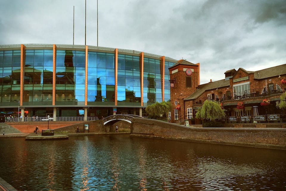 A modern building with glass windows reflecting the sky, a brick structure with a sign, and a canal with a bridge.