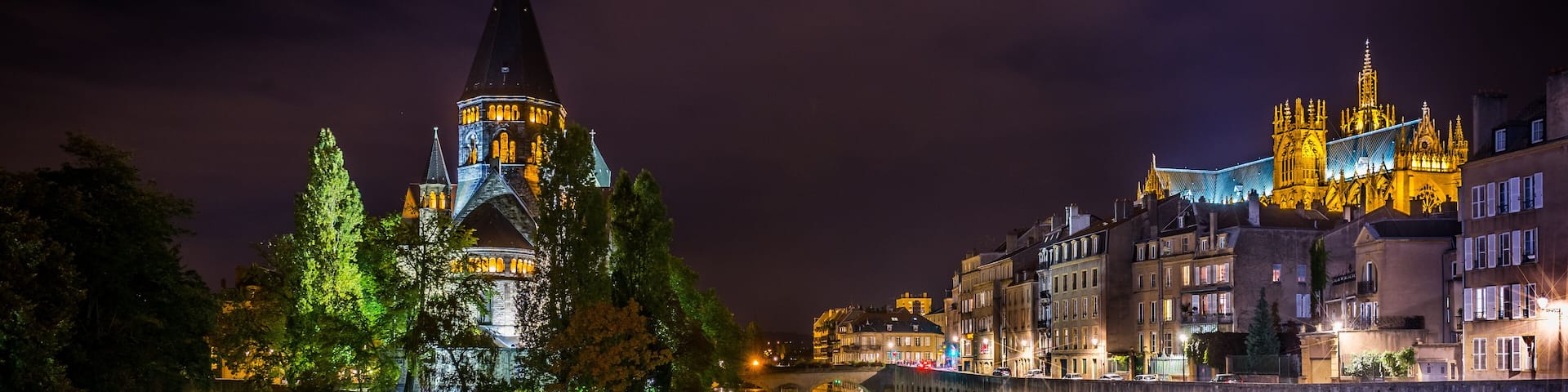 A nighttime view of a castle with a spire, reflecting in a calm river, surrounded by illuminated buildings.