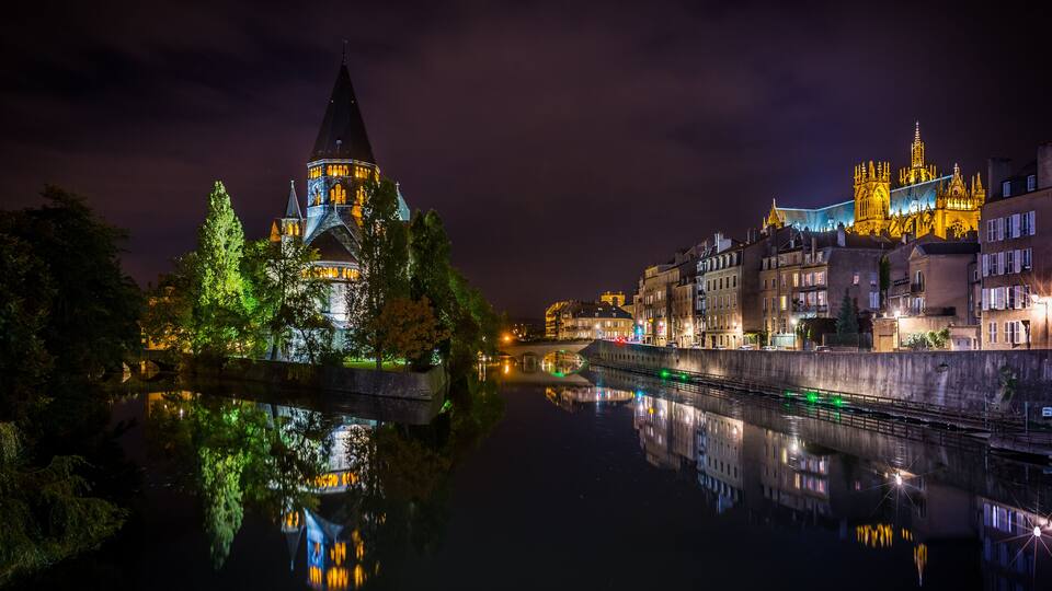 A nighttime view of a castle with a spire, reflecting in a calm river, surrounded by illuminated buildings.
