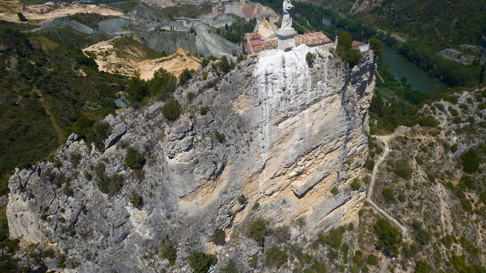 A rocky cliff with a statue on top, a river below, and a distant town.