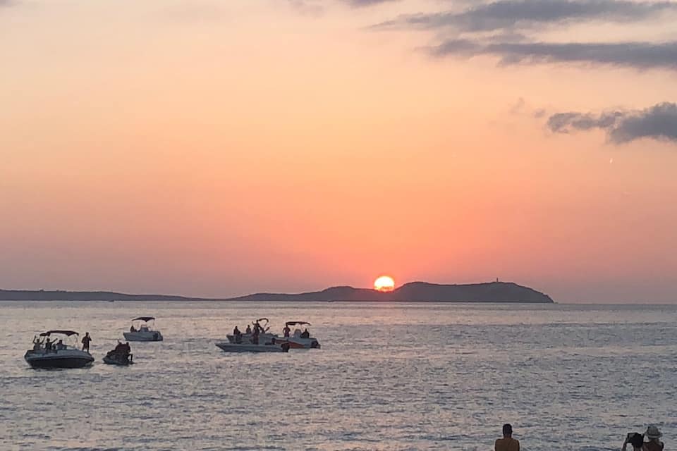 Un coucher de soleil sur une mer calme, avec plusieurs bateaux et une île lointaine.