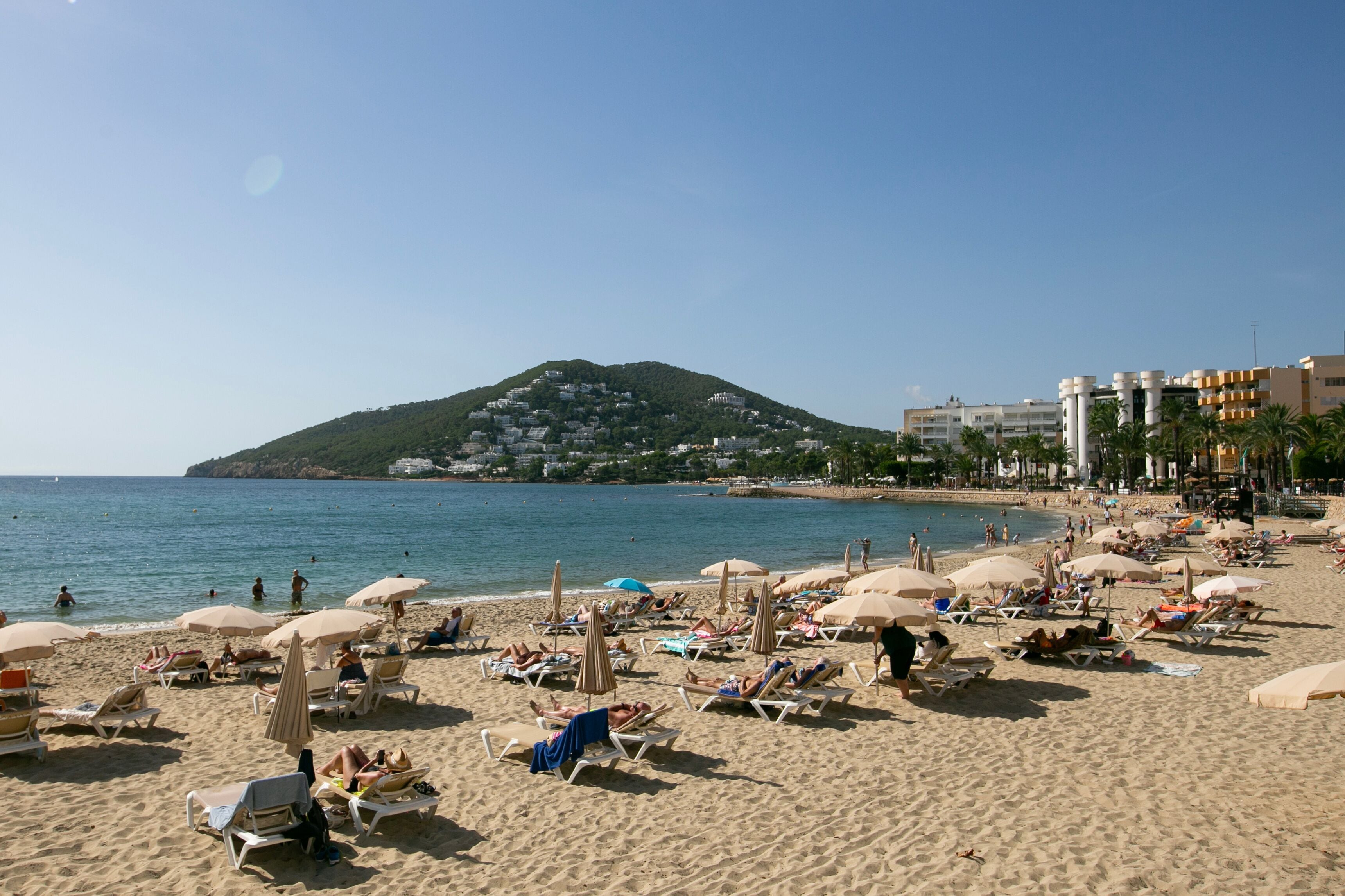 A beach with sun loungers and umbrellas, a clear sky, and a hill in the background.