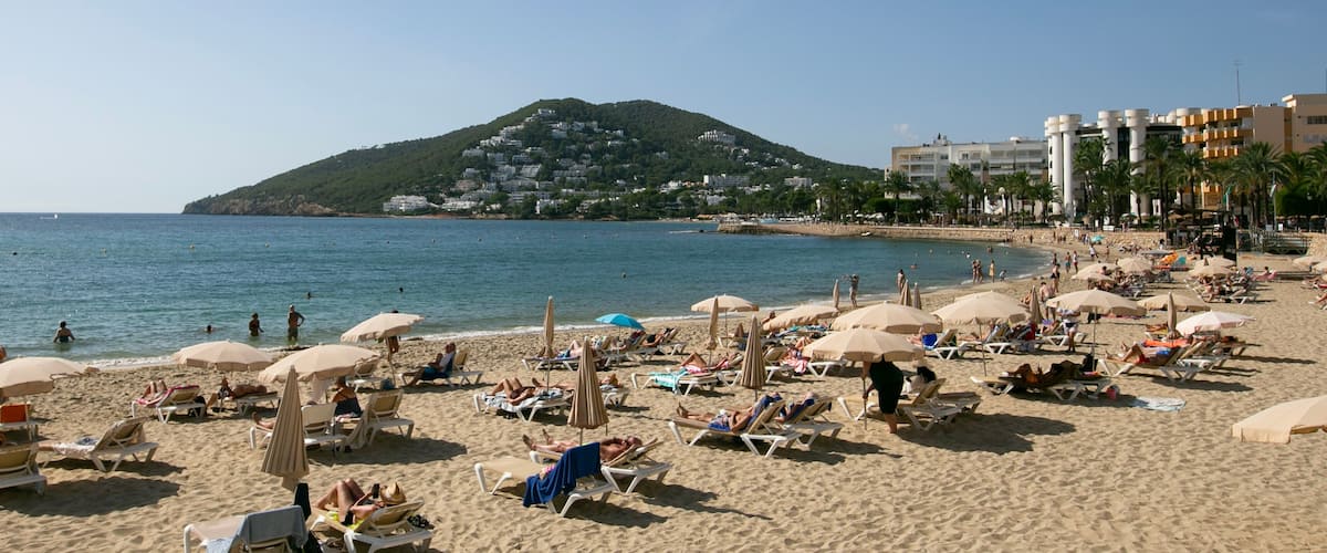 A beach with sun loungers and umbrellas, a clear sky, and a hill in the background.