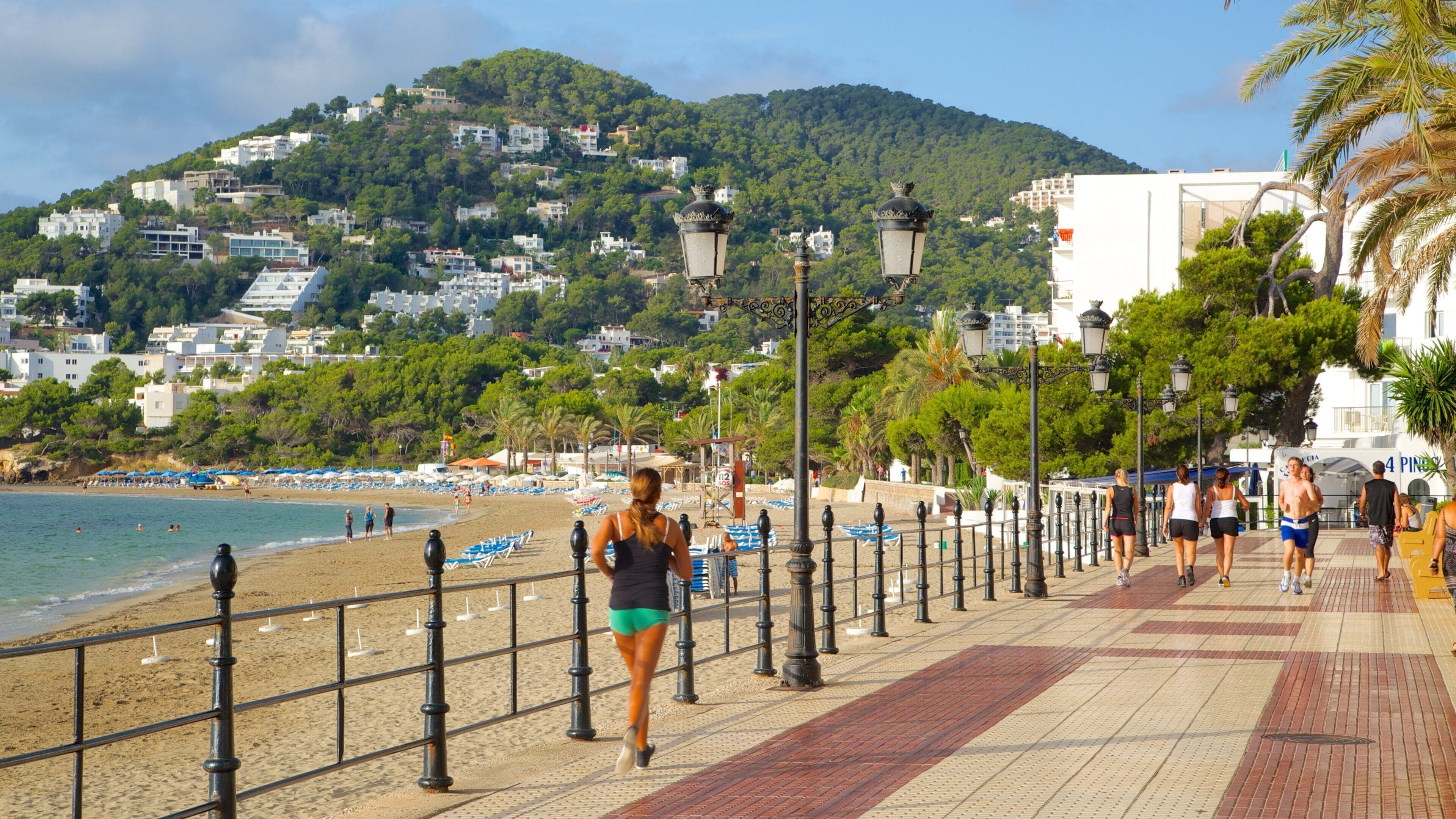 Un paseo marítimo frente a una playa, con sombrillas y montañas al fondo.
