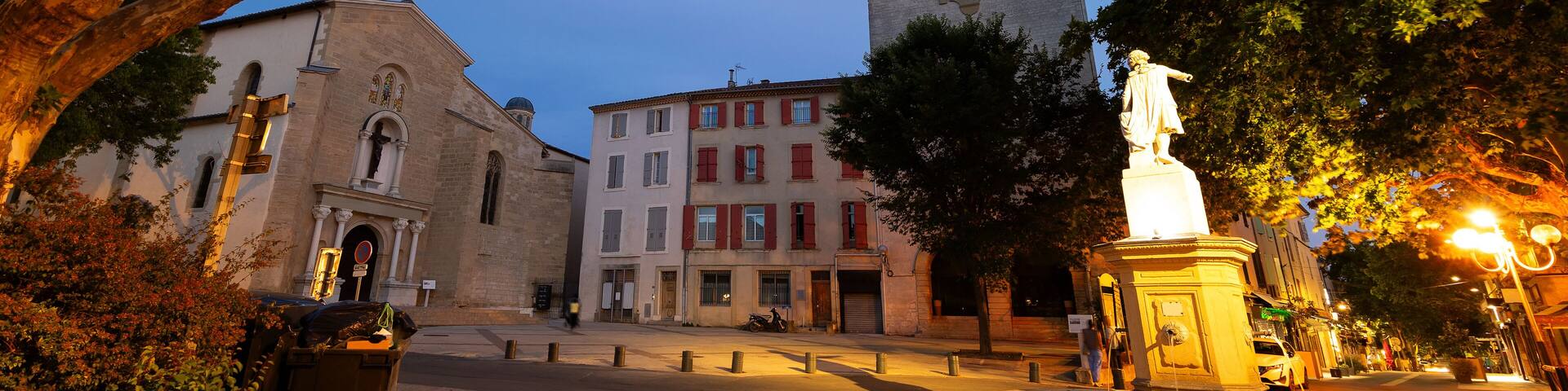 Historic square Place Mirabeau in French town of Pertuis overlooking illuminated statue with fountain on background with medieval donjon with clock and parish church of Saint-Nicolas on summer evening