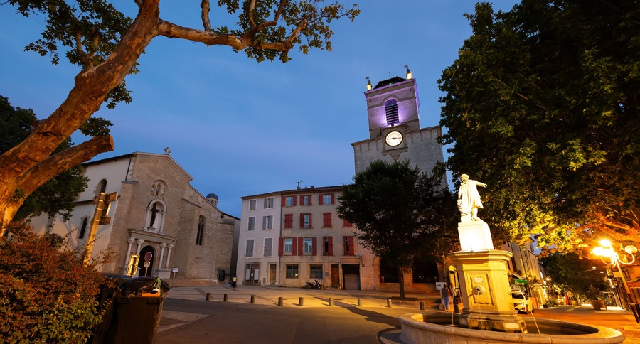 Historic square Place Mirabeau in French town of Pertuis overlooking illuminated statue with fountain on background with medieval donjon with clock and parish church of Saint-Nicolas on summer evening