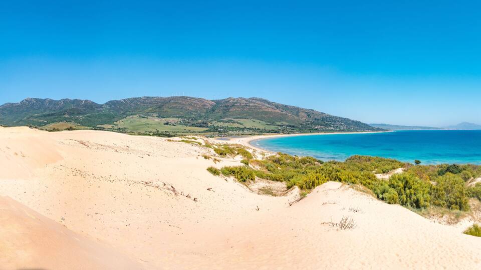 A coastal landscape with sand dunes, a beach, and a mountain range in the background.