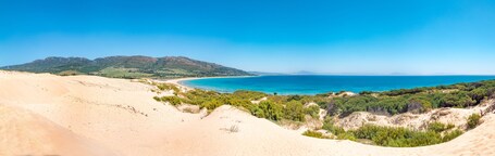 Eine Küstenlandschaft mit Dünen, einem Strand und einem Gebirgszug im Hintergrund.