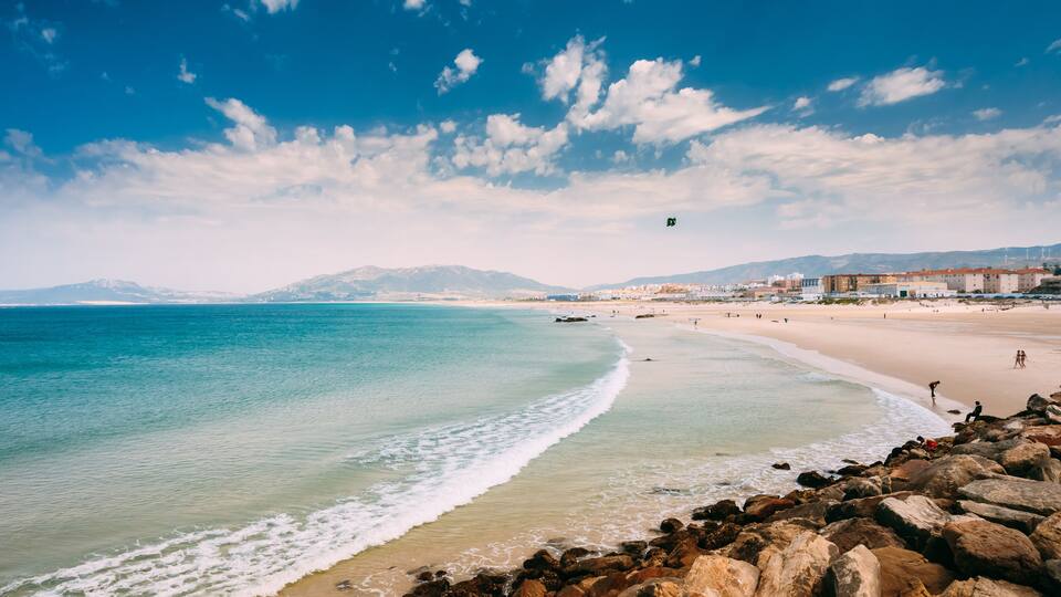 A coastal beach with rocky shoreline, clear blue water, and distant mountains under a partly cloudy sky.