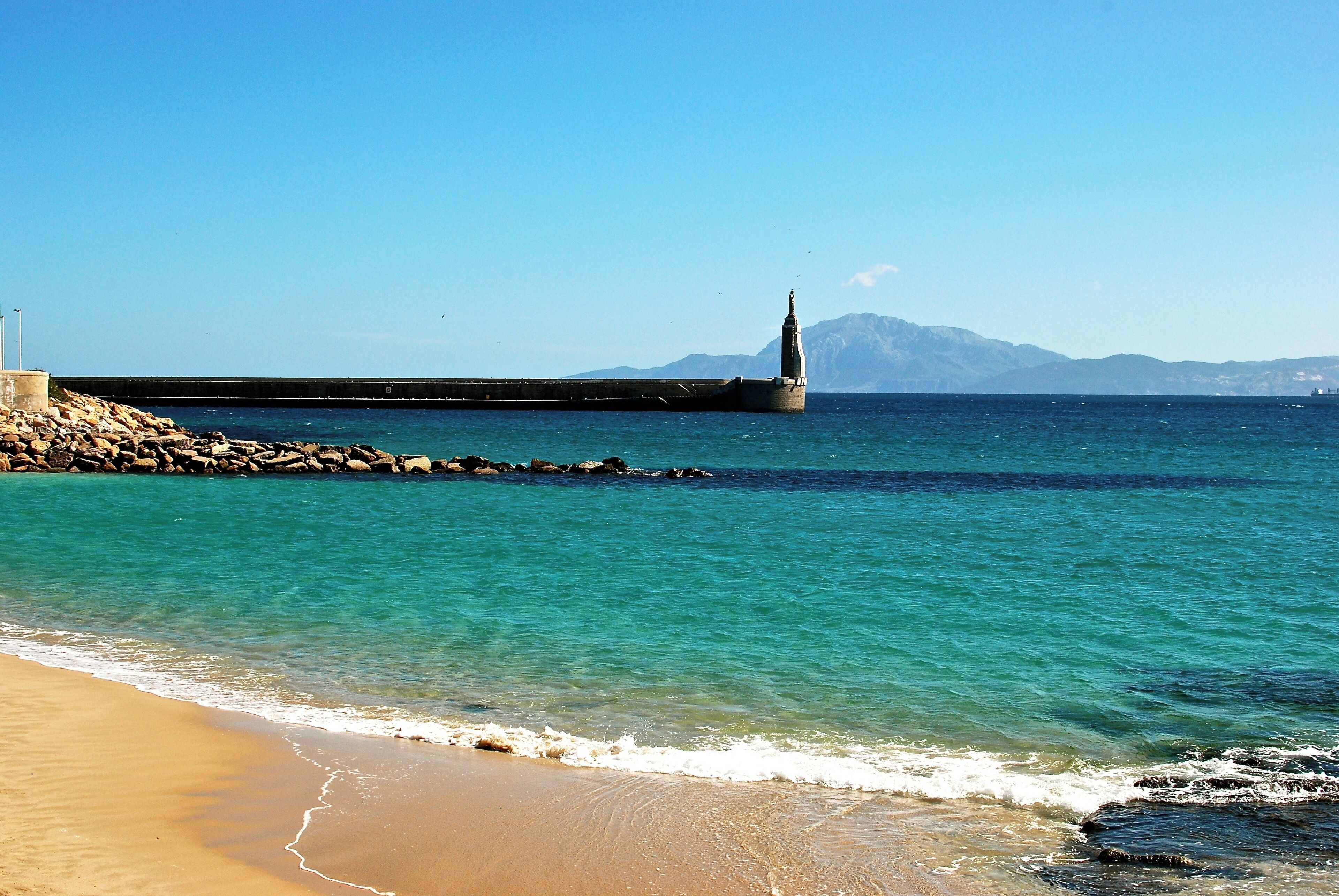 A beach with clear turquoise water, a rocky breakwater, and a distant mountain range.