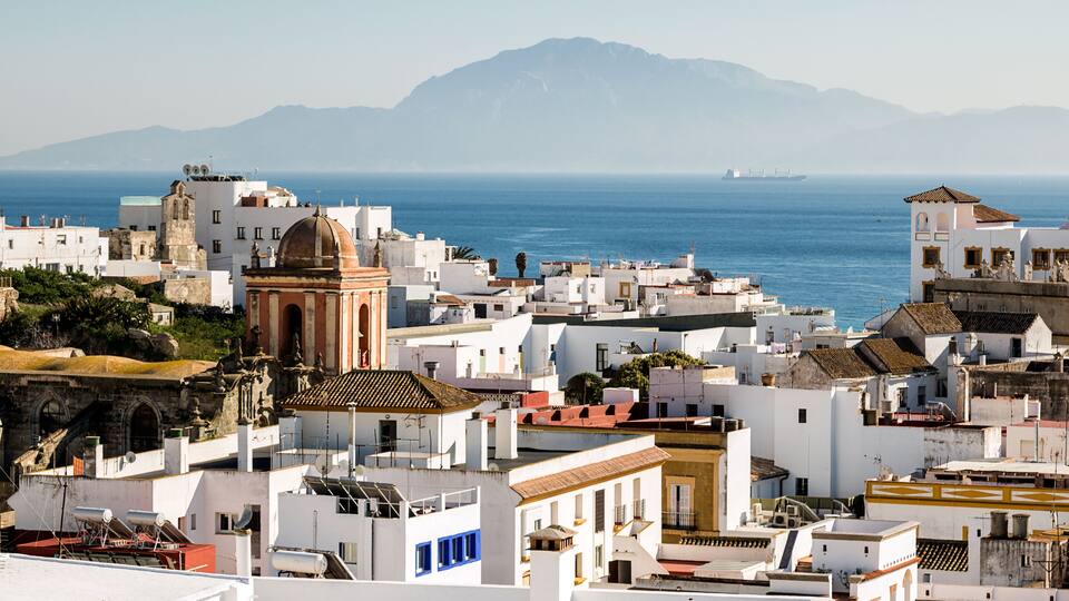 A coastal cityscape with white buildings, a prominent church, and a mountain range in the background.