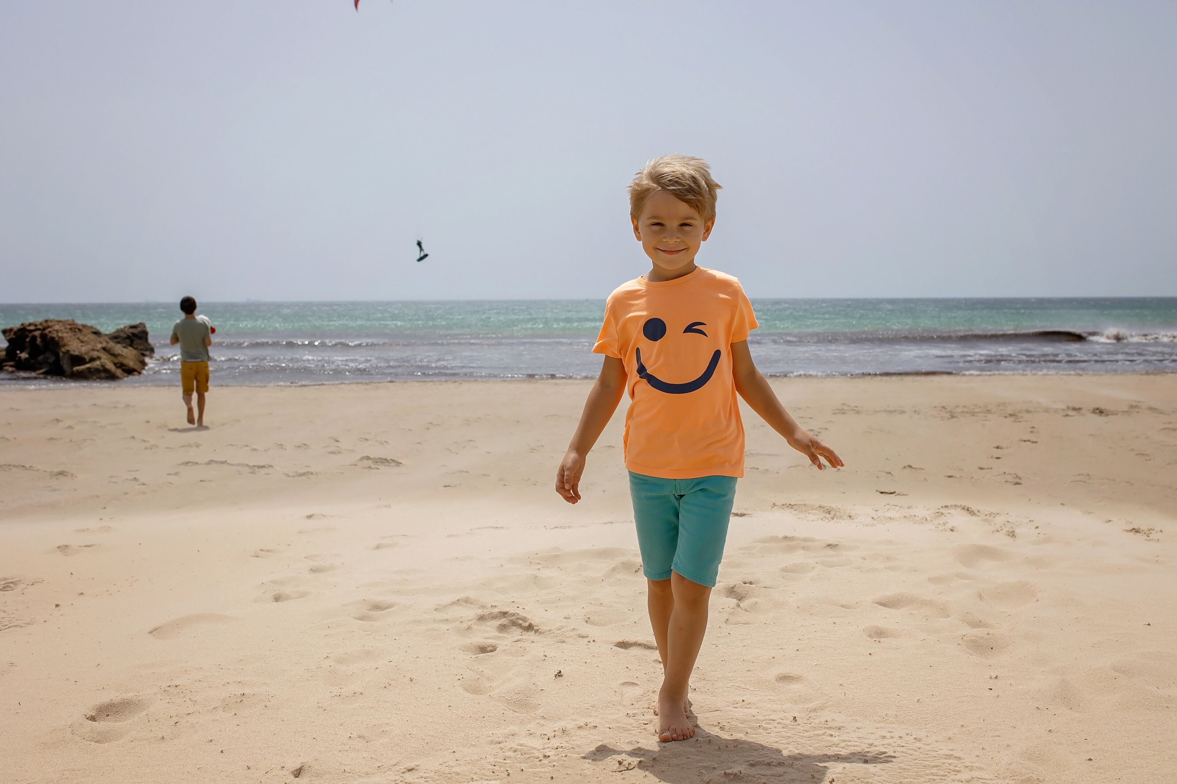Niño caminando sobre una playa arenosa con un mar tranquilo y rocas al fondo.