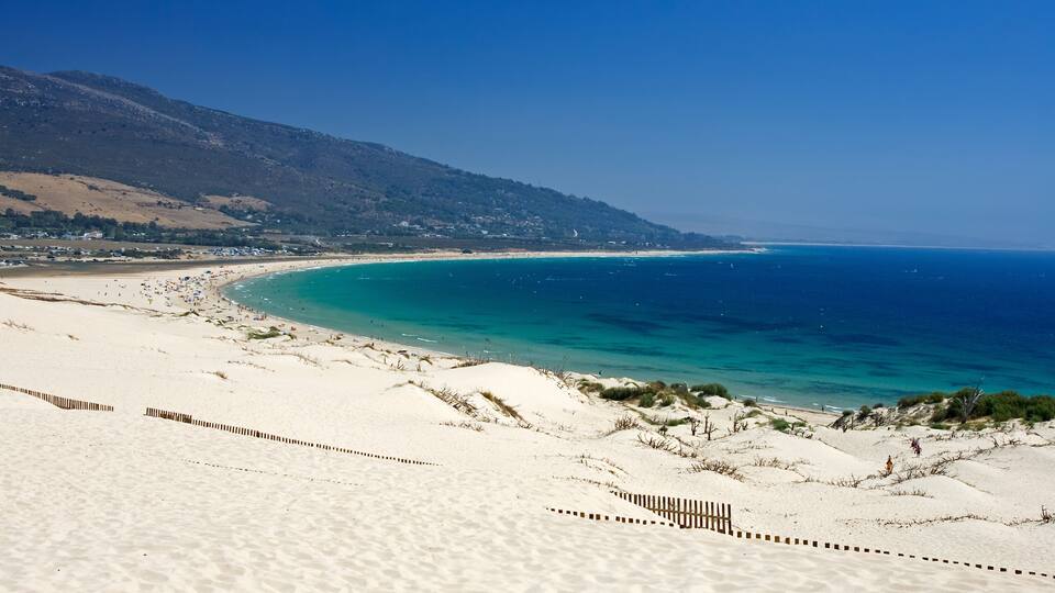 A beach with white sand, clear turquoise water, and a mountainous backdrop.