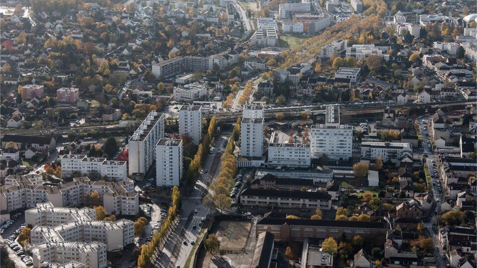 An aerial view of a city with dense residential buildings, a prominent road, and surrounding greenery.