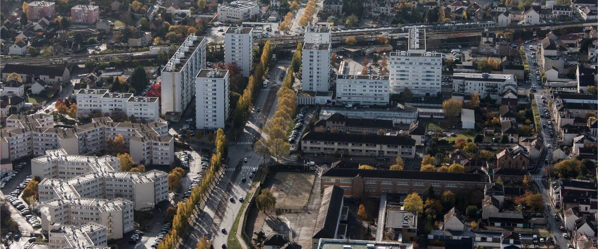 An aerial view of a city with dense residential buildings, a prominent road, and surrounding greenery.