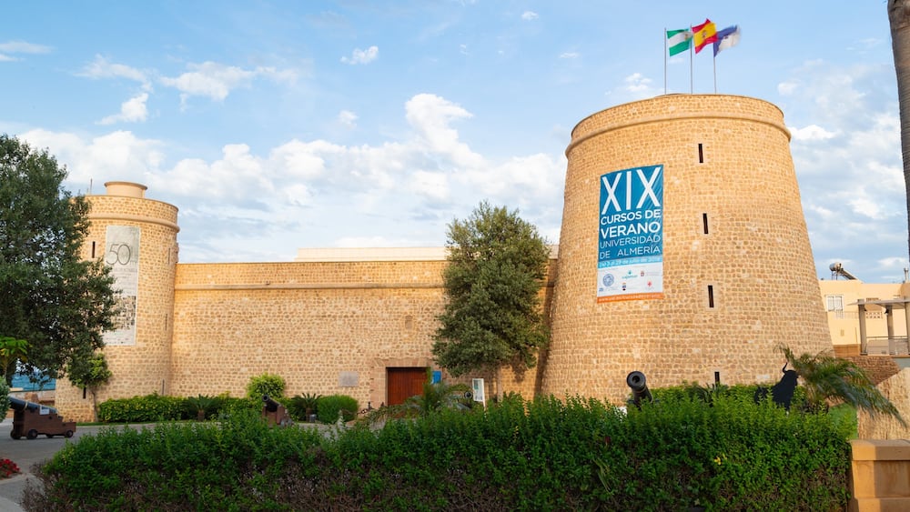 Un bâtiment historique en pierre, orné d’un drapeau, d’un canon et entouré d’une végétation luxuriante.