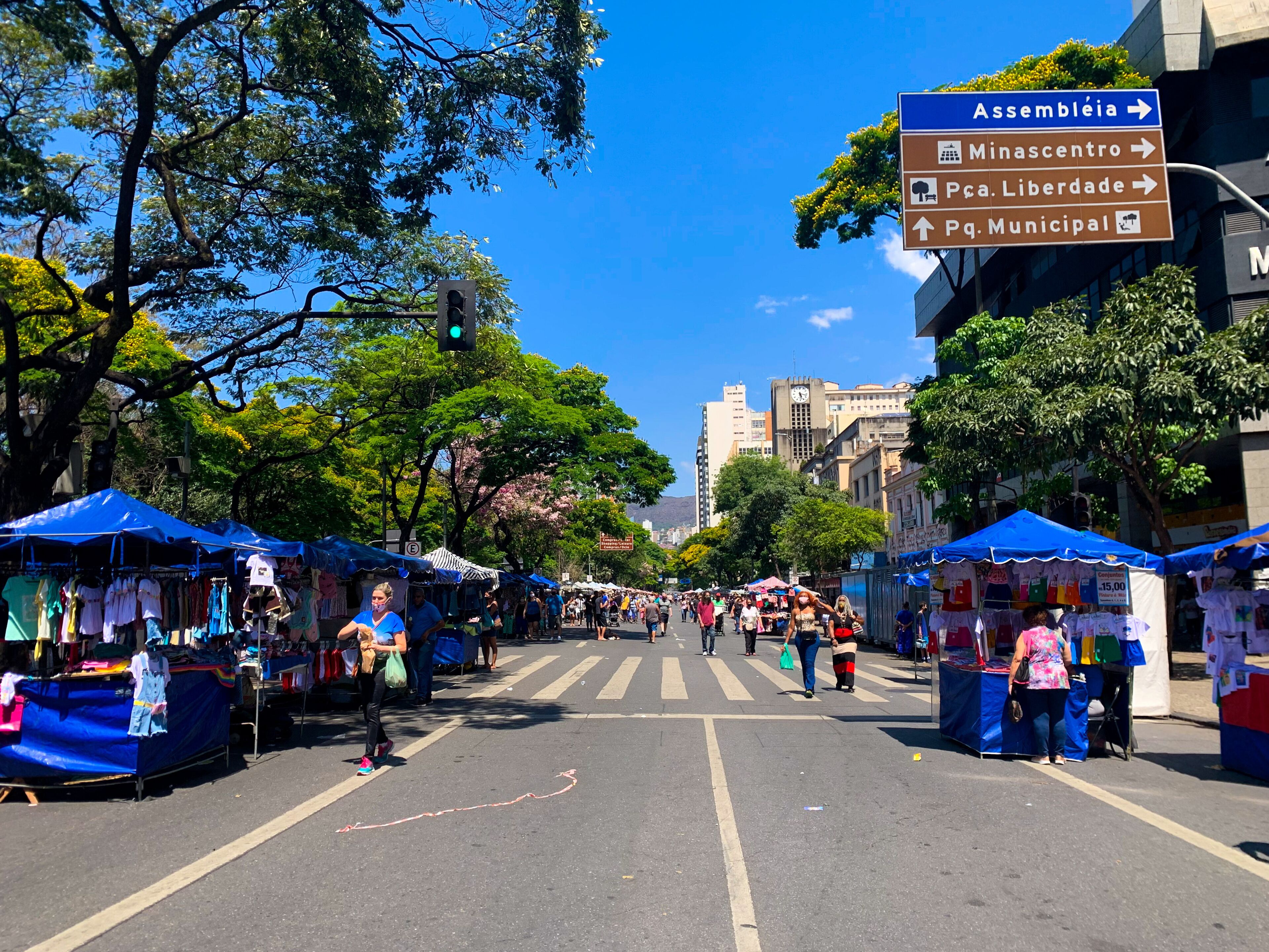 A street market with vendors under blue canopies, a pedestrian crossing, and directional signs for Assembledia, Minascentro, Pça. Libertade, and Pq. Municipal.