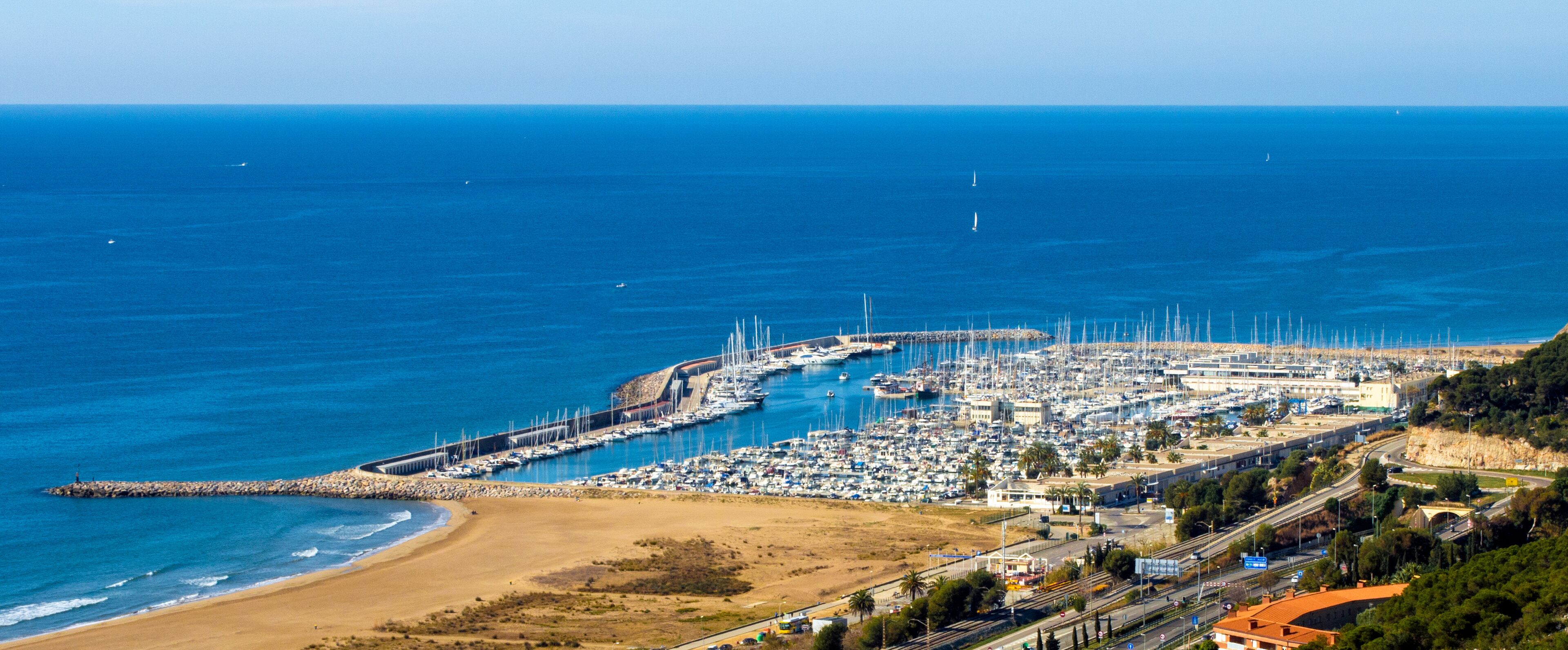 A coastal marina with numerous boats, a sandy beach, and a rocky breakwater.