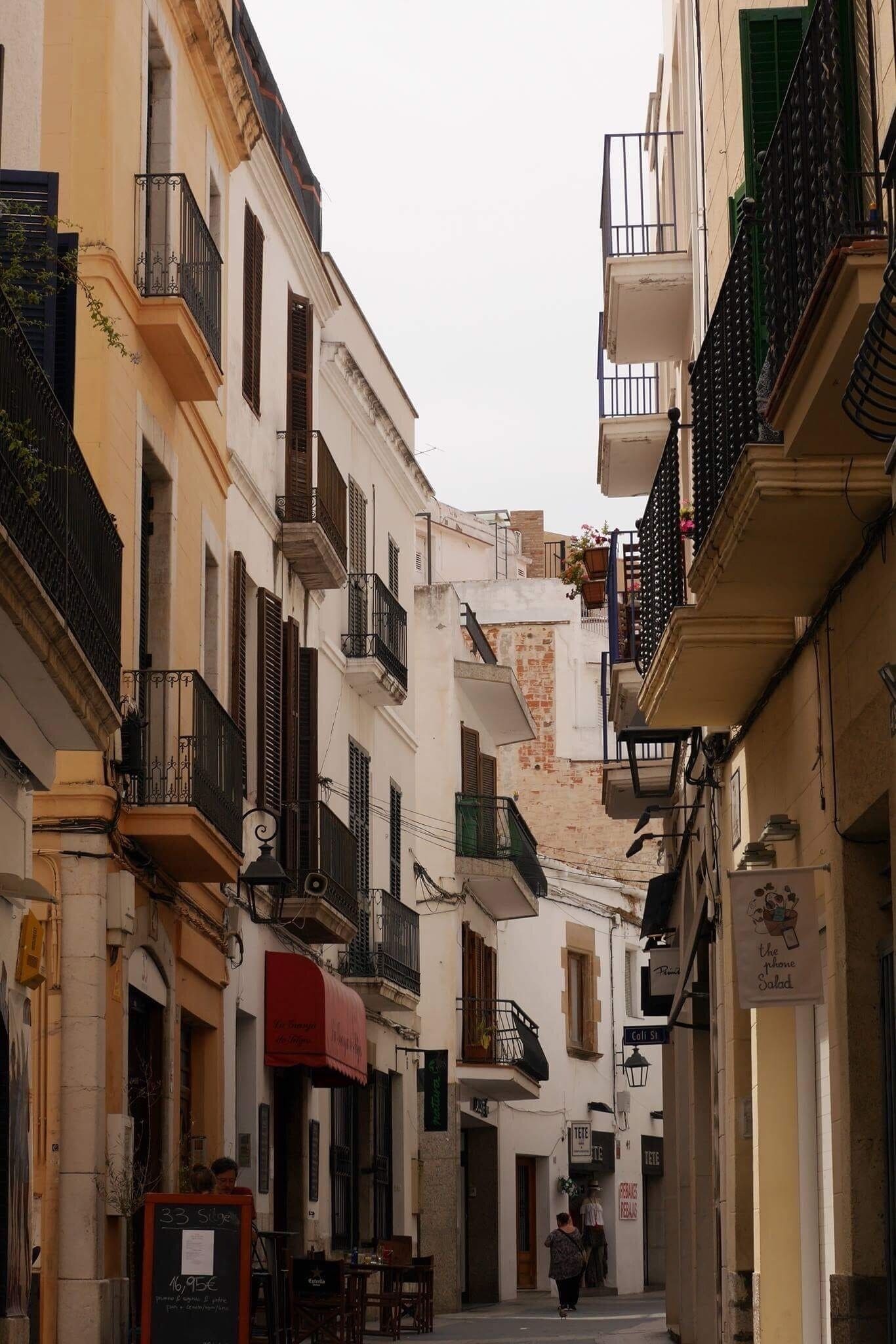 Uma rua estreita com edifícios de ambos os lados, com varandas, toldos e placas.