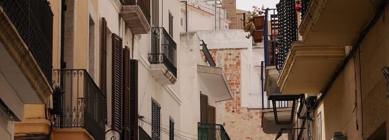 Uma rua estreita com edifícios de ambos os lados, com varandas, toldos e placas.