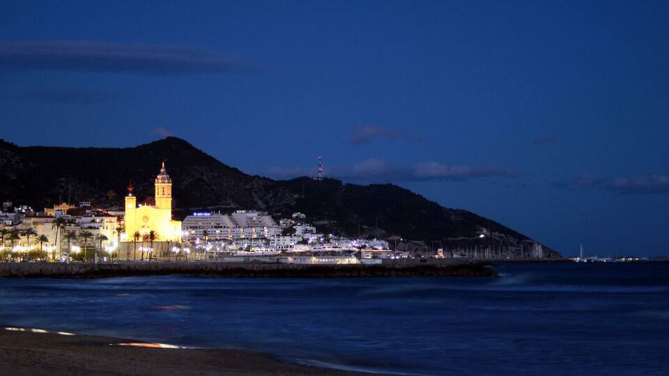 A coastal town at night with illuminated buildings, a prominent church, and a mountain in the background.