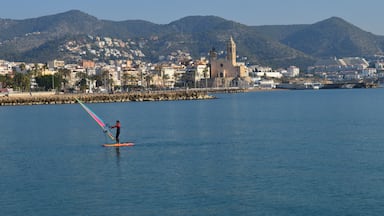 Una persona practica windsurf en un mar tranquilo, con un pueblo costero y una iglesia al fondo.