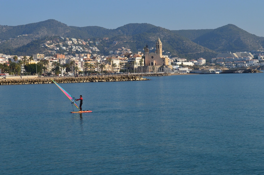Una persona practica windsurf en un mar tranquilo, con un pueblo costero y una iglesia al fondo.