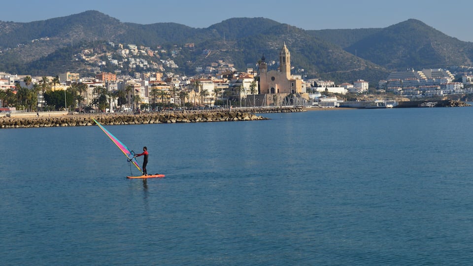 A person windsurfing on a calm sea with a coastal town and a church in the background.
