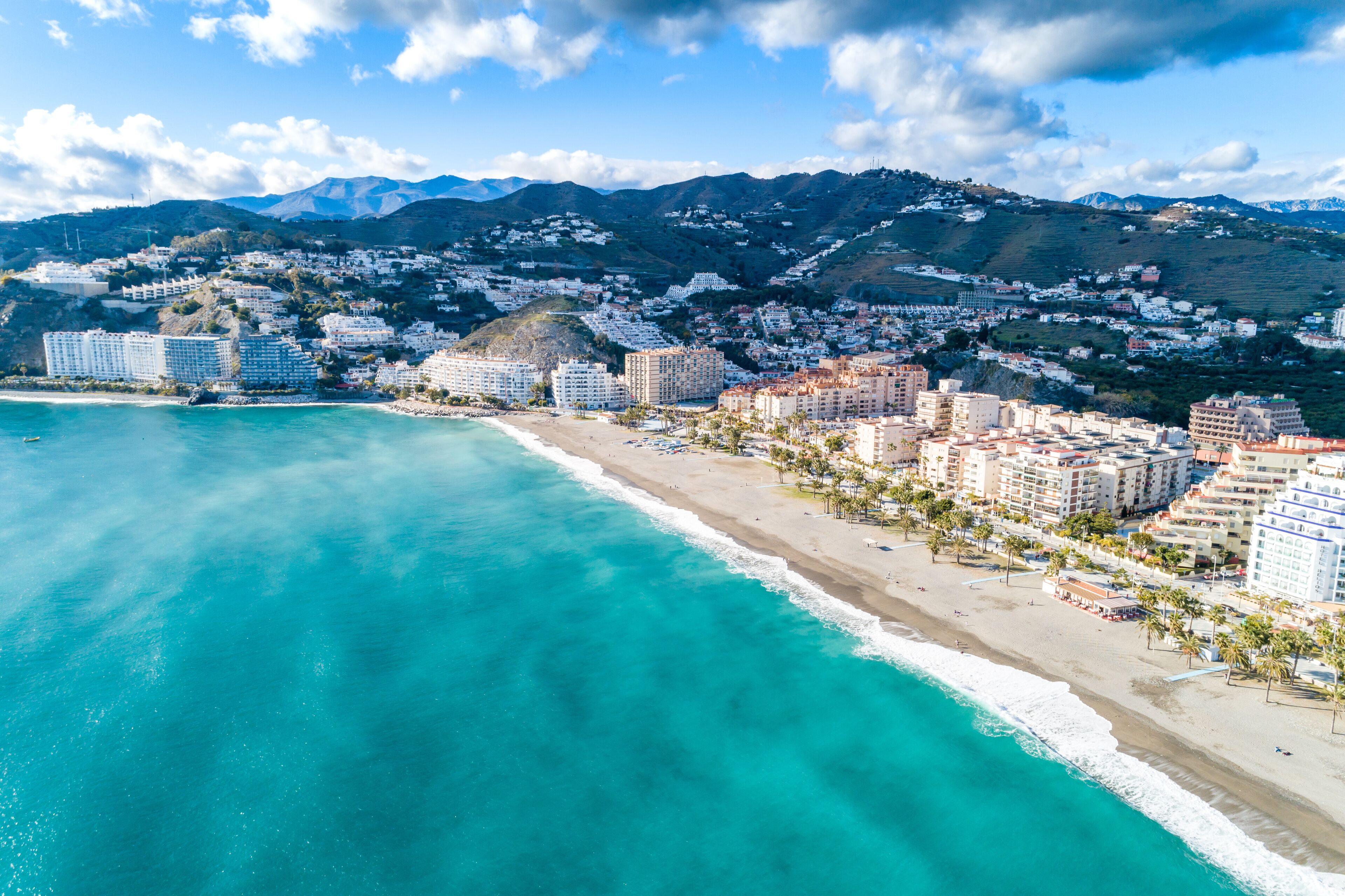 A coastal cityscape with a beach, buildings, and mountains in the background.