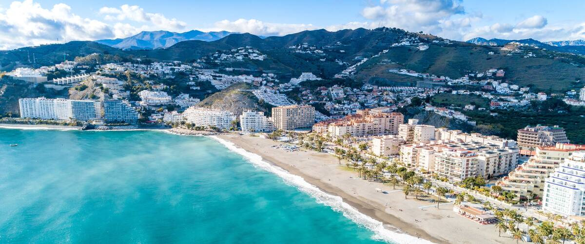 A coastal cityscape with a beach, buildings, and mountains in the background.