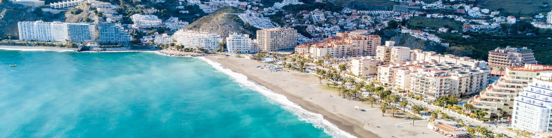 A coastal cityscape with a beach, buildings, and mountains in the background.