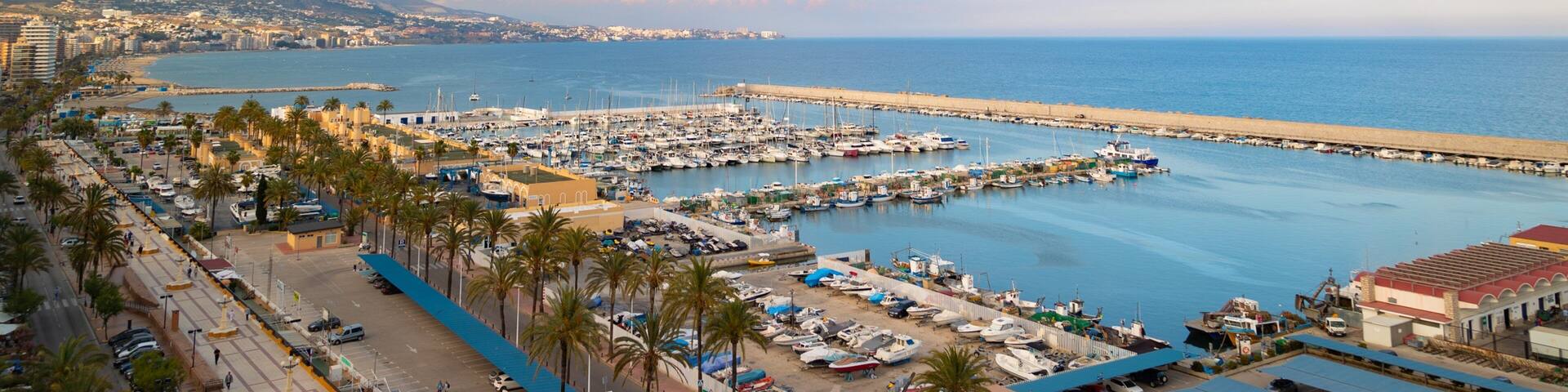A coastal marina with boats, a beach, and a mountainous backdrop.