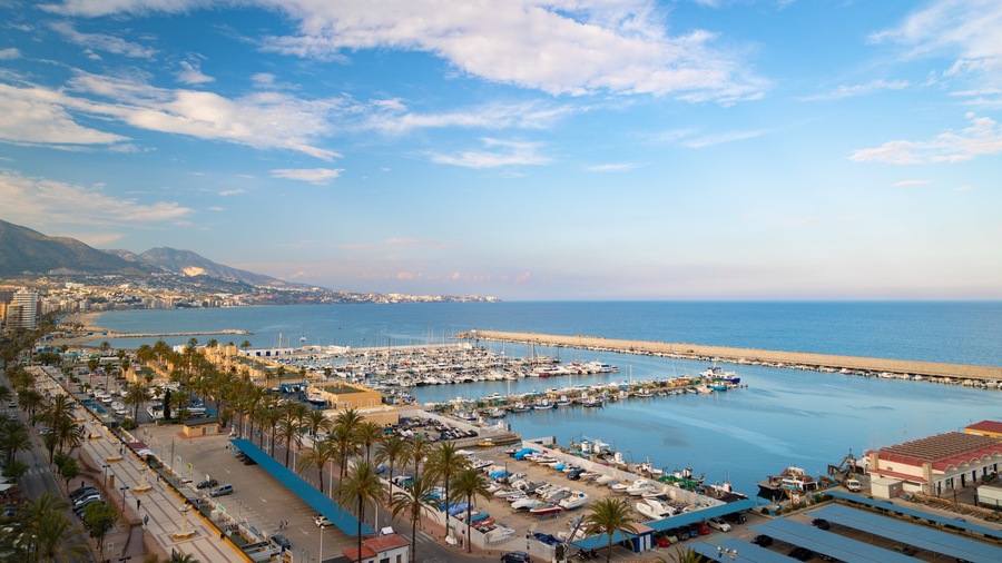 A coastal marina with boats, a beach, and a mountainous backdrop.