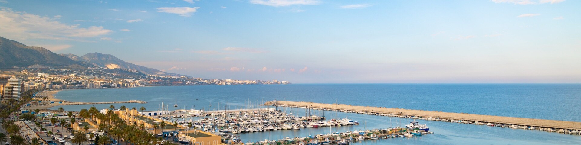 A coastal marina with boats, a beach, and a mountainous backdrop.