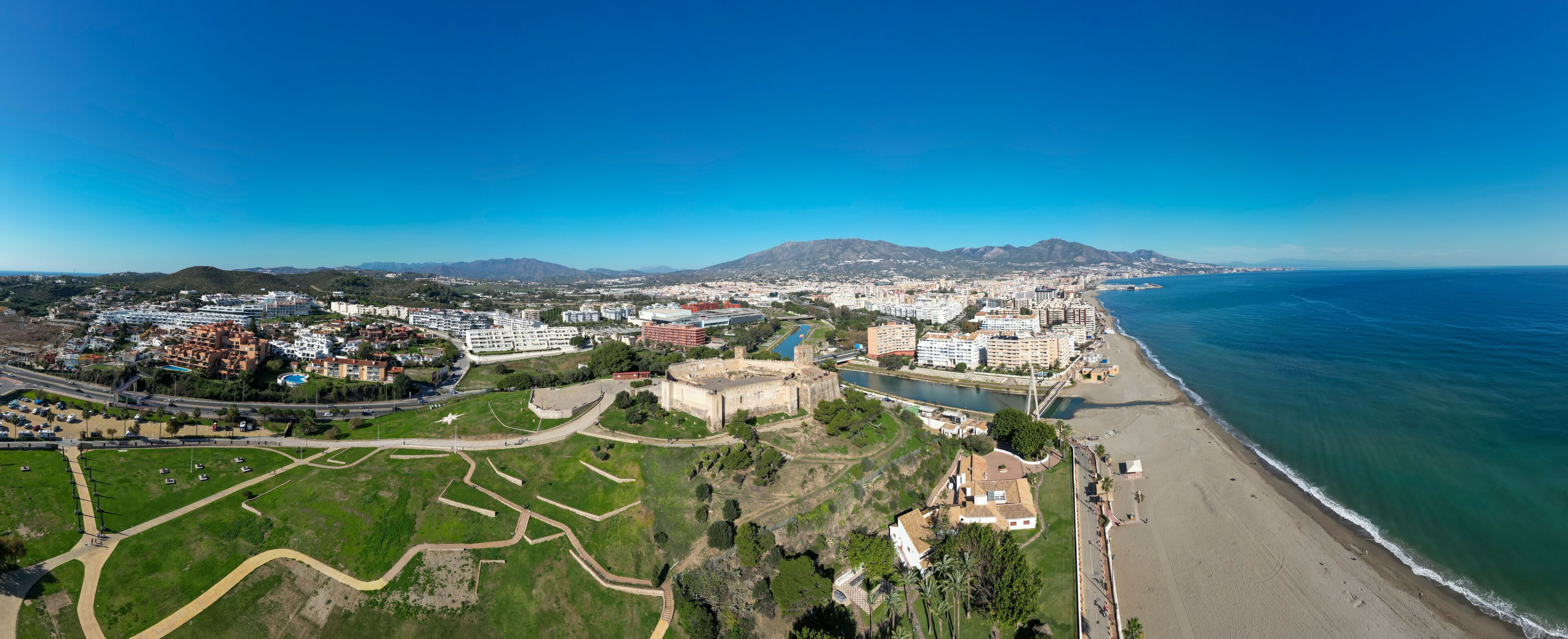 A coastal cityscape with a prominent castle, a beach, and a mountain range in the background.