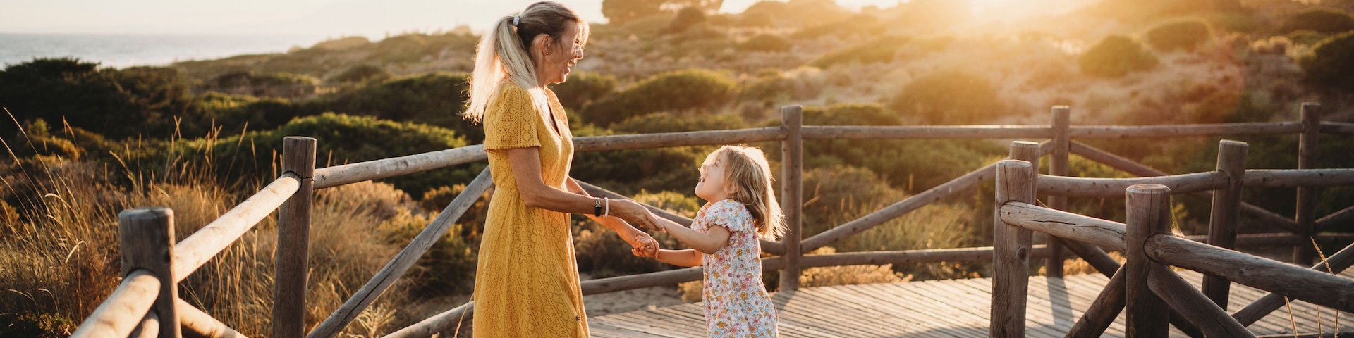 Pasarela de madera con una mujer y un niño tomados de la mano, con vistas a un paisaje costero con dunas y vegetación.