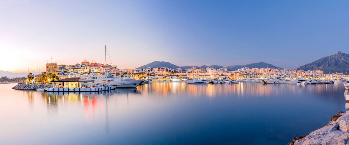 A coastal town with a marina, buildings, and a mountain in the background.