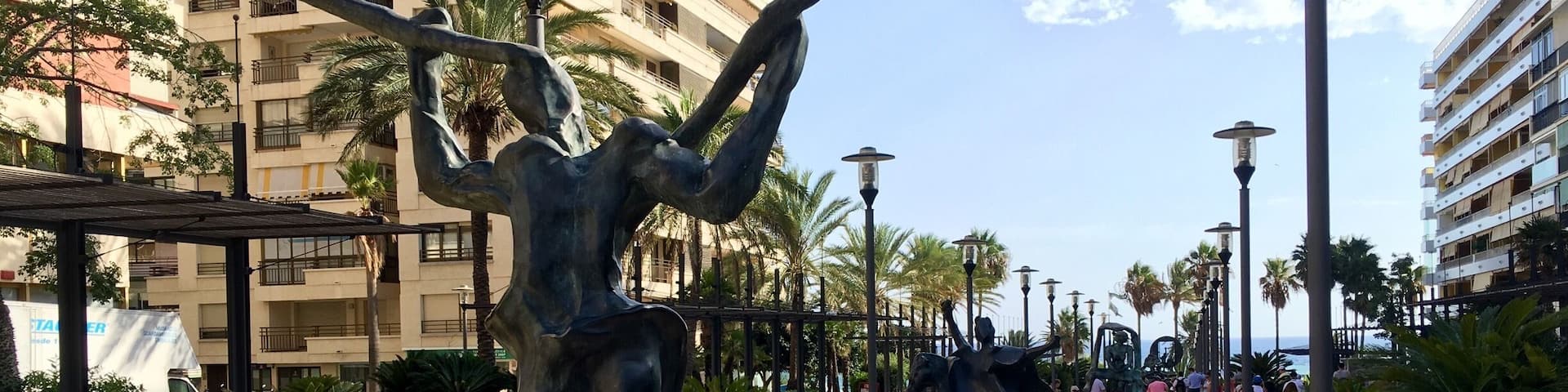 Una plaza pública con esculturas, palmeras y un edificio al fondo.