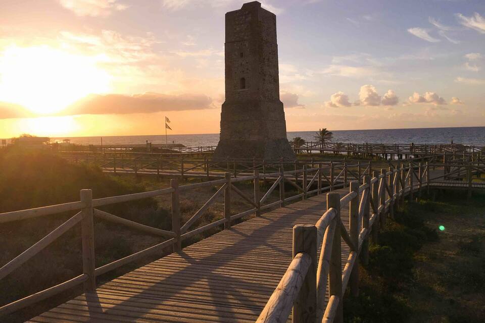 A wooden boardwalk leading to a tall stone tower by the sea during sunset.