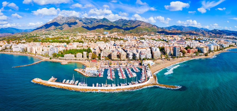 A coastal city with a marina, mountains in the background, and a clear sky.