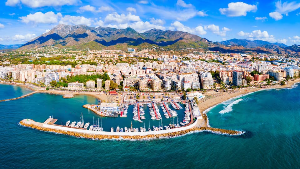 A coastal city with a marina, mountains in the background, and a clear sky.