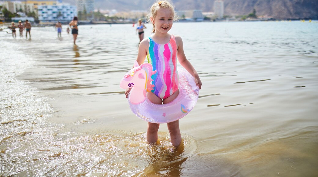 Little girl in a colorful swimsuit standing in shallow sea water with an inflatable unicorn swim ring in Los Cristianos, Tenerife, Canary Islands, Spain.