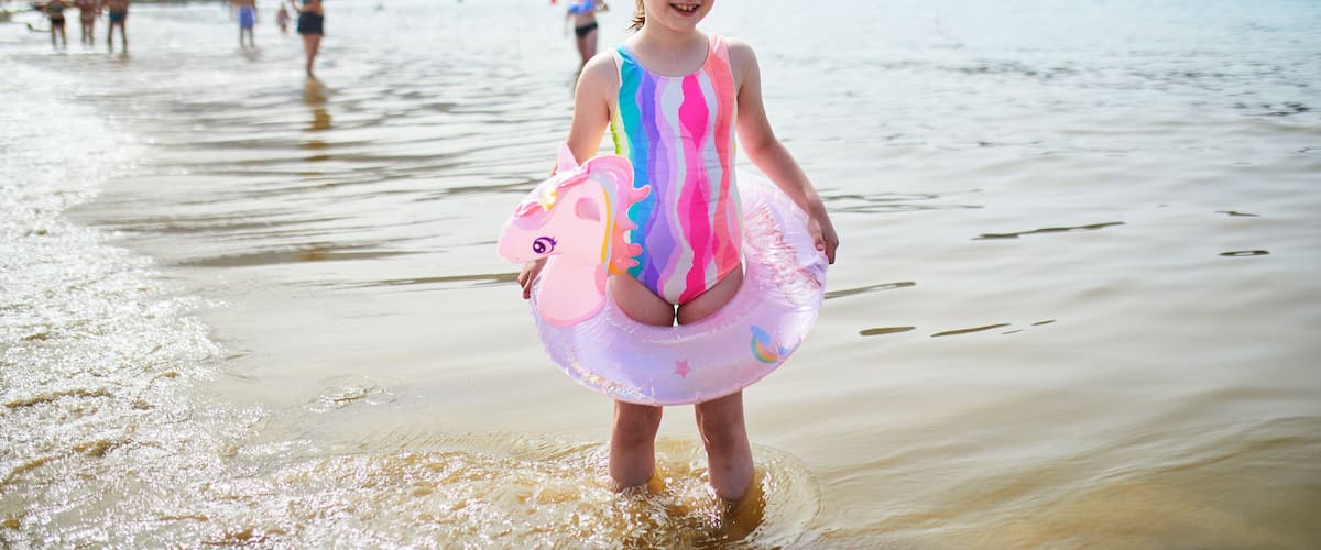 A beach with clear water, a child in a colorful swimsuit, and buildings in the background.