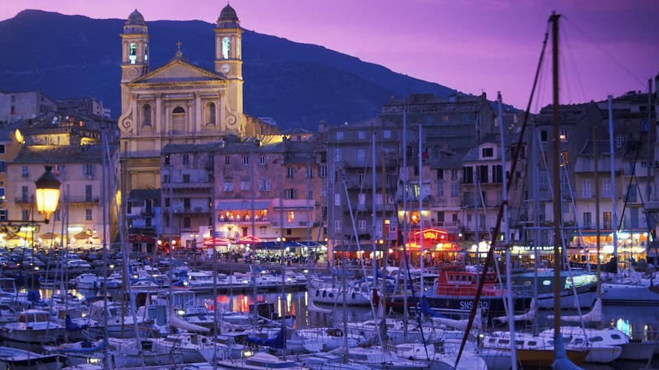 A marina with boats, a church with two towers, and buildings with lights against a purple sky.