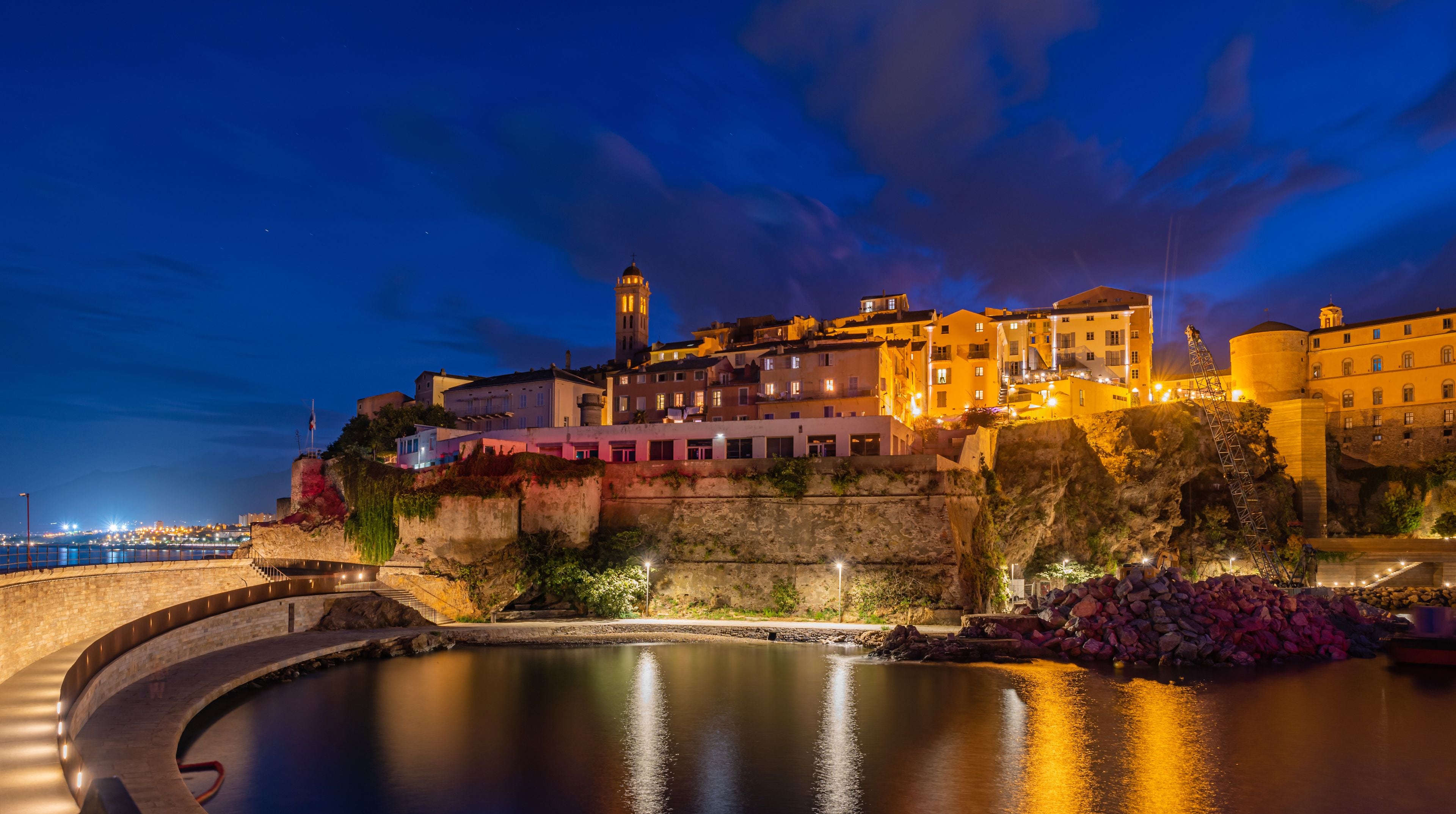 A nighttime coastal cityscape with illuminated buildings, a prominent tower, and a calm body of water reflecting the lights.