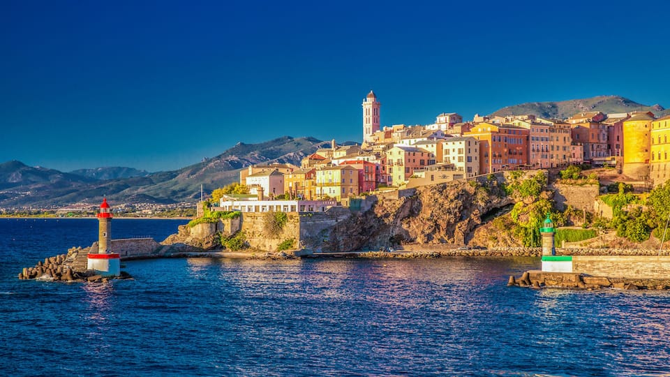 A coastal town with colorful buildings, a lighthouse, and mountains in the background.