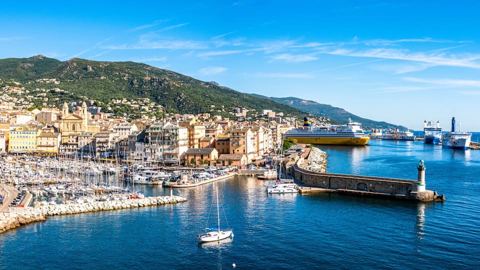 A coastal town with a marina, boats, a lighthouse, and a mountain in the background.