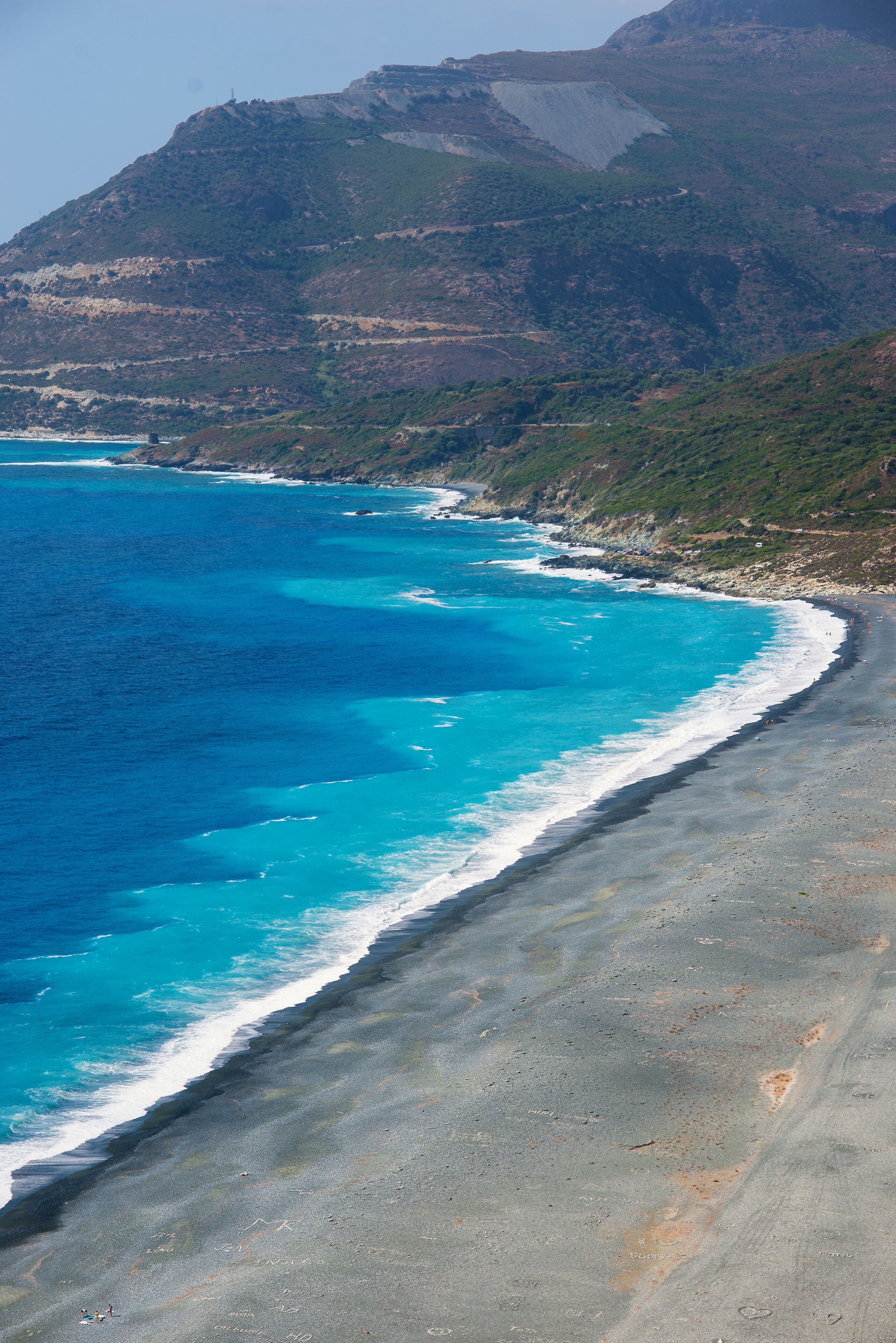 Un paisaje costero con mar azul transparente, una playa de arena y montañas al fondo.
