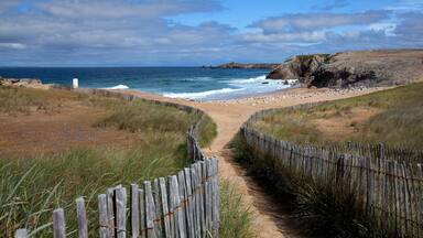 Un sendero costero que lleva a una playa arenosa con una valla de madera, acantilados rocosos y el océano.