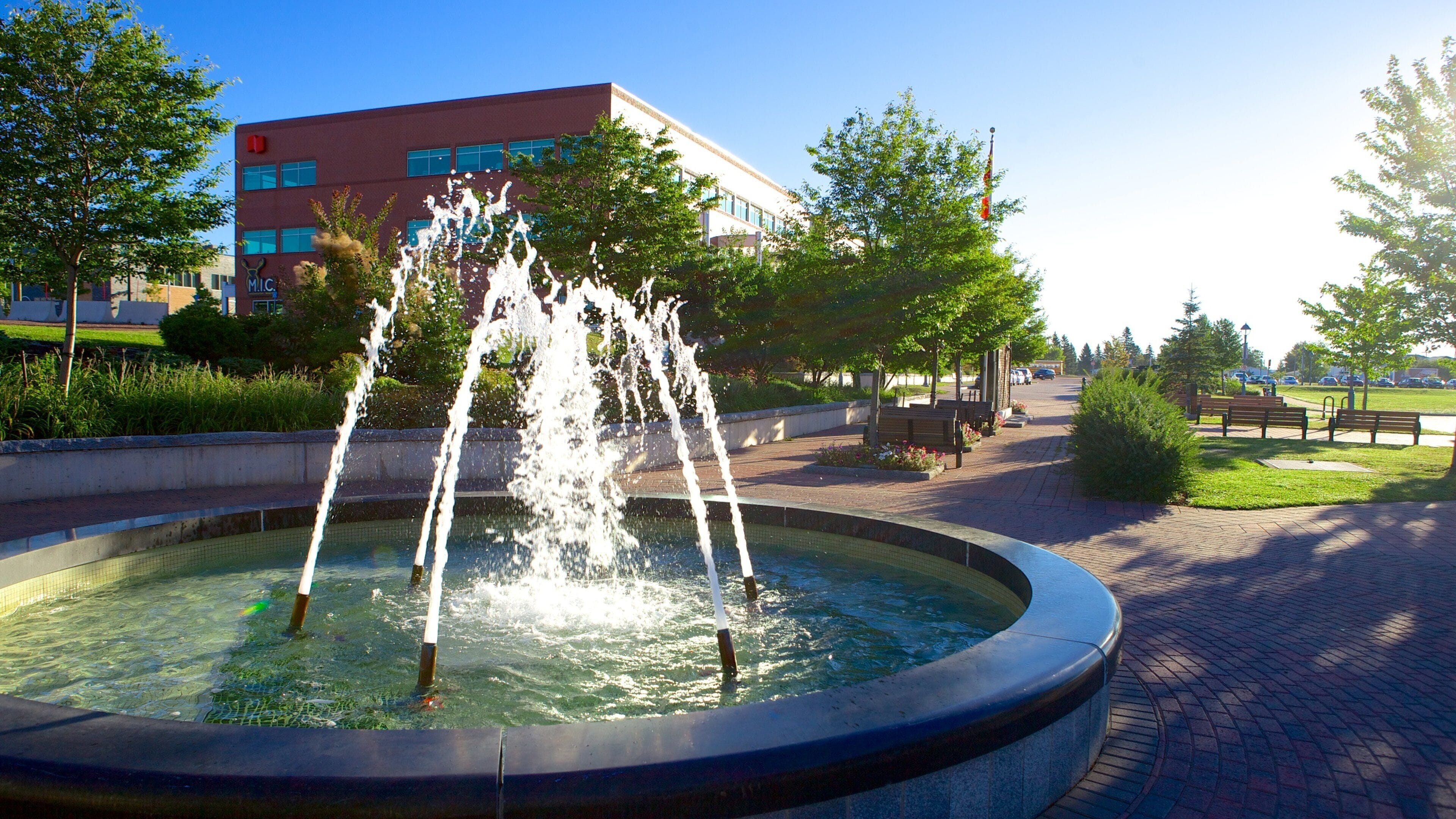 A fountain with water jets in a circular basin, surrounded by a paved walkway, benches, and a building in the background.